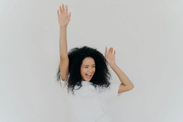 Joyful woman with curly hair raising hands, smiling against a plain white background