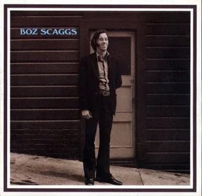 A man in a suit stands on a sidewalk beside a door and dark-paneled wall; the text "Boz Scaggs" appears in the top left corner.