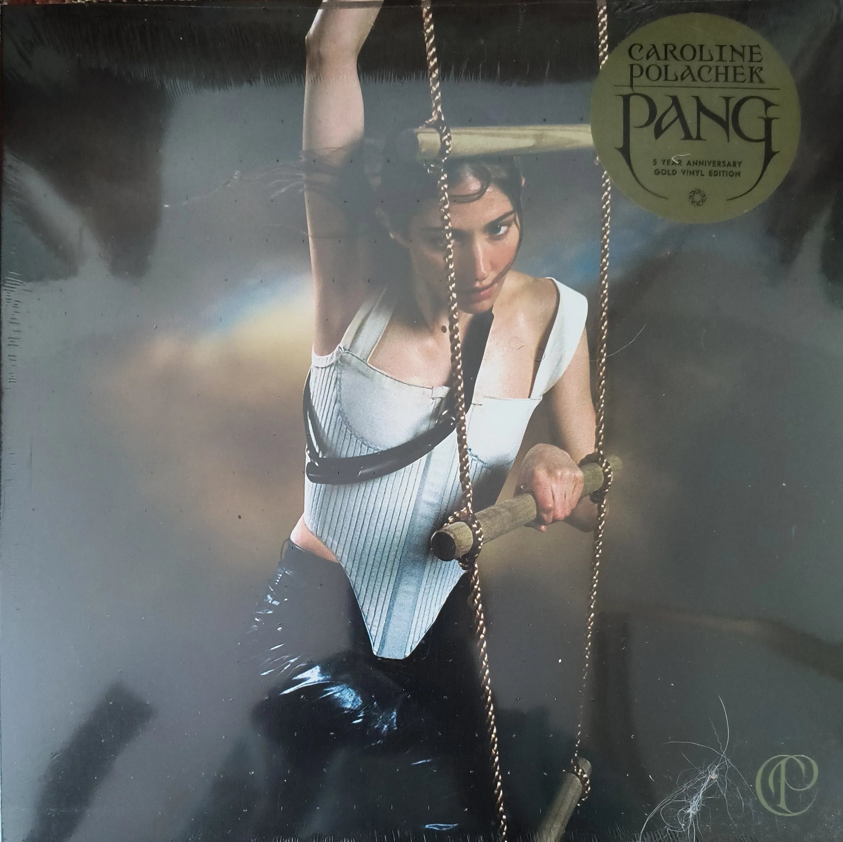 A woman in a white top and black pants holds wooden swings. The album cover displays "Caroline Polachek PANG 5th Anniversary Gold Vinyl Edition" in a gold sticker on the top right.