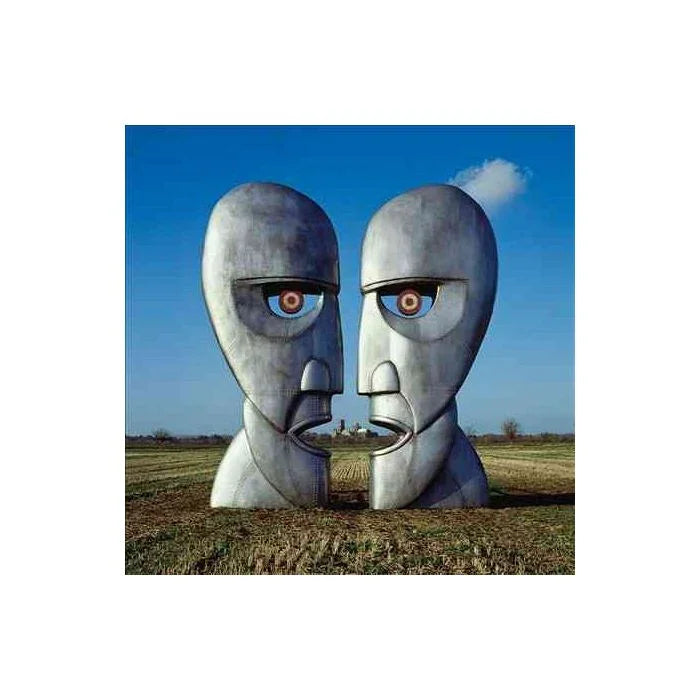 Two large metallic face sculptures positioned to face each other, forming a third face in the negative space between them, set in an open field under a blue sky.