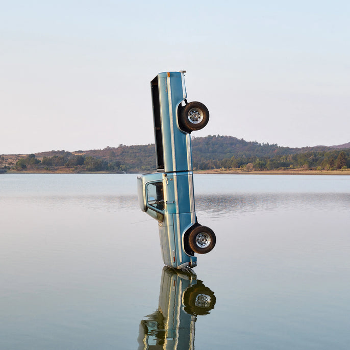 Blue pickup truck standing vertically in a lake with hills in the background, artistic cover photo.