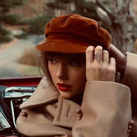 A person wearing a brown hat and beige coat sits in a car, touching the brim of their hat with one hand and looking to the side.