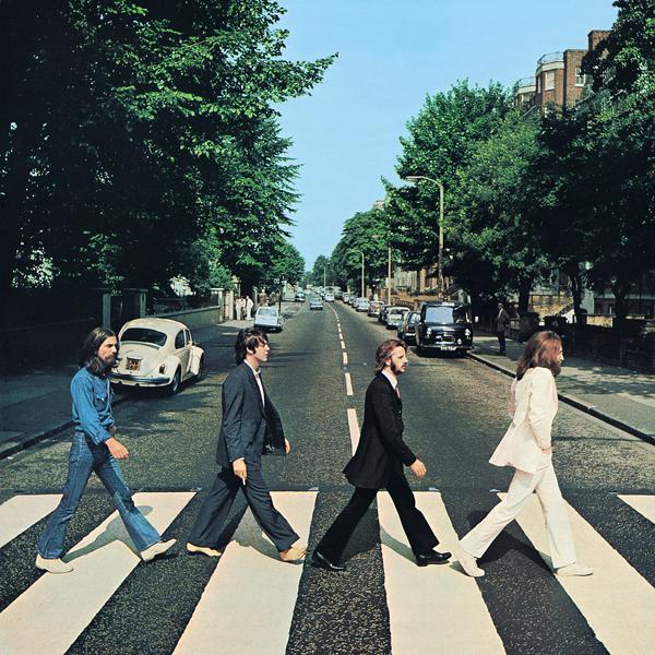 Four men walk across a zebra crossing on a tree-lined street with cars parked along the side, under a clear blue sky.