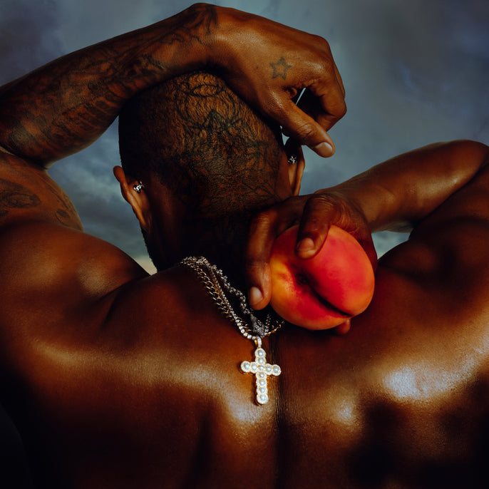 Tattooed man holding peach behind back, wearing cross necklace, dramatic lighting