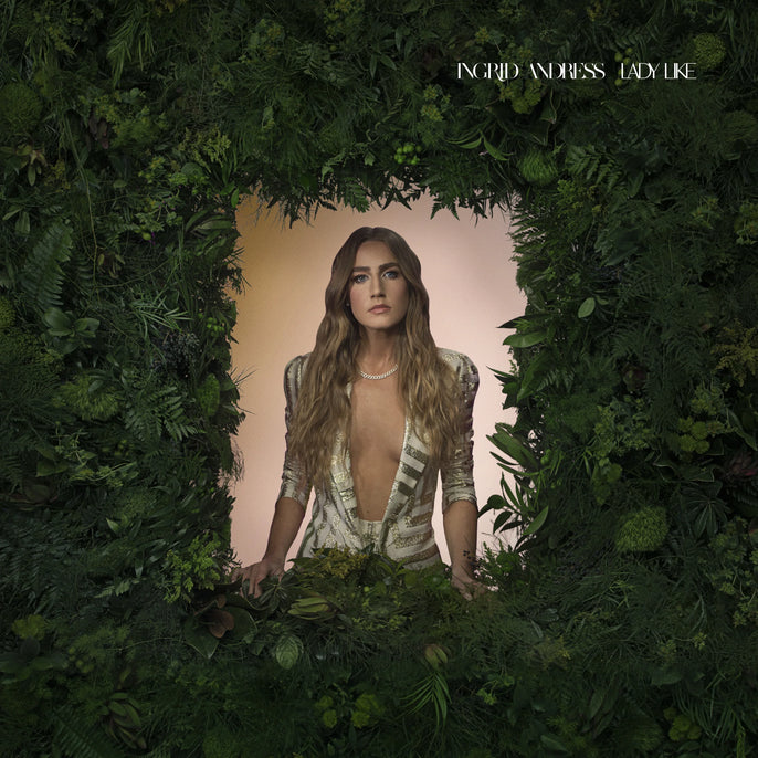 A woman with long hair stands at a window framed with dense green foliage, wearing a deep V-neck dress; text in the corner reads "Ingrid Andress Lady Like.