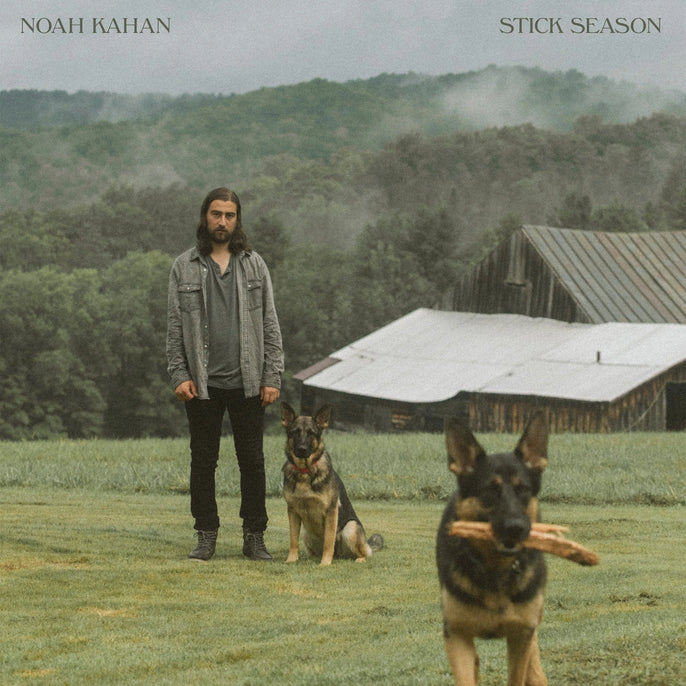 A man stands on grass near two German Shepherds; one dog holds sticks in its mouth. A wooden barn and misty trees are in the background. The text reads "Noah Kahan Stick Season.