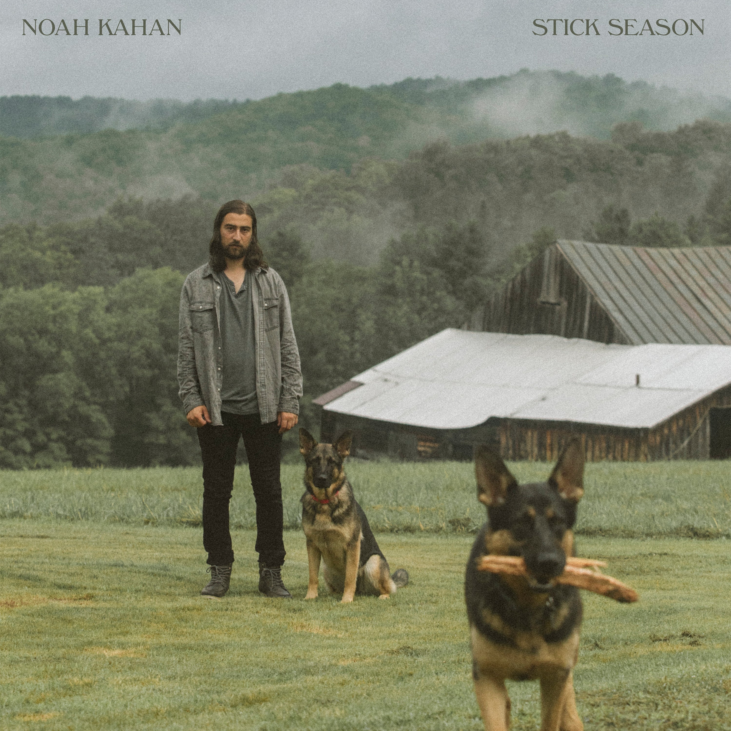 A man stands on grass near two German Shepherds; one dog holds sticks in its mouth. A wooden barn and misty trees are in the background. The text reads "Noah Kahan Stick Season.