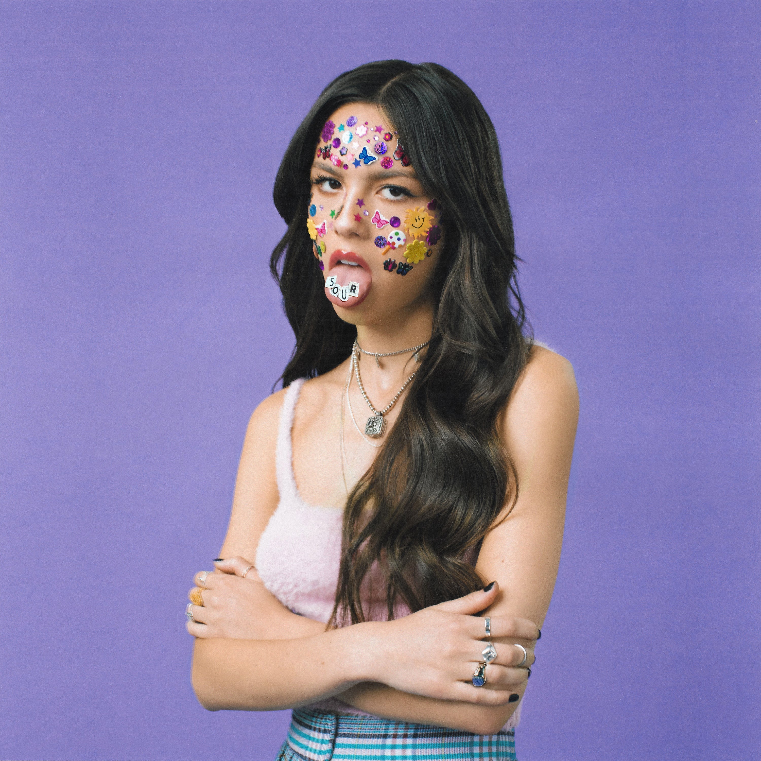 A young woman with long dark hair stands against a purple background, arms crossed, face covered in colorful stickers, and a white sticker reading "SOUR" on her tongue.
