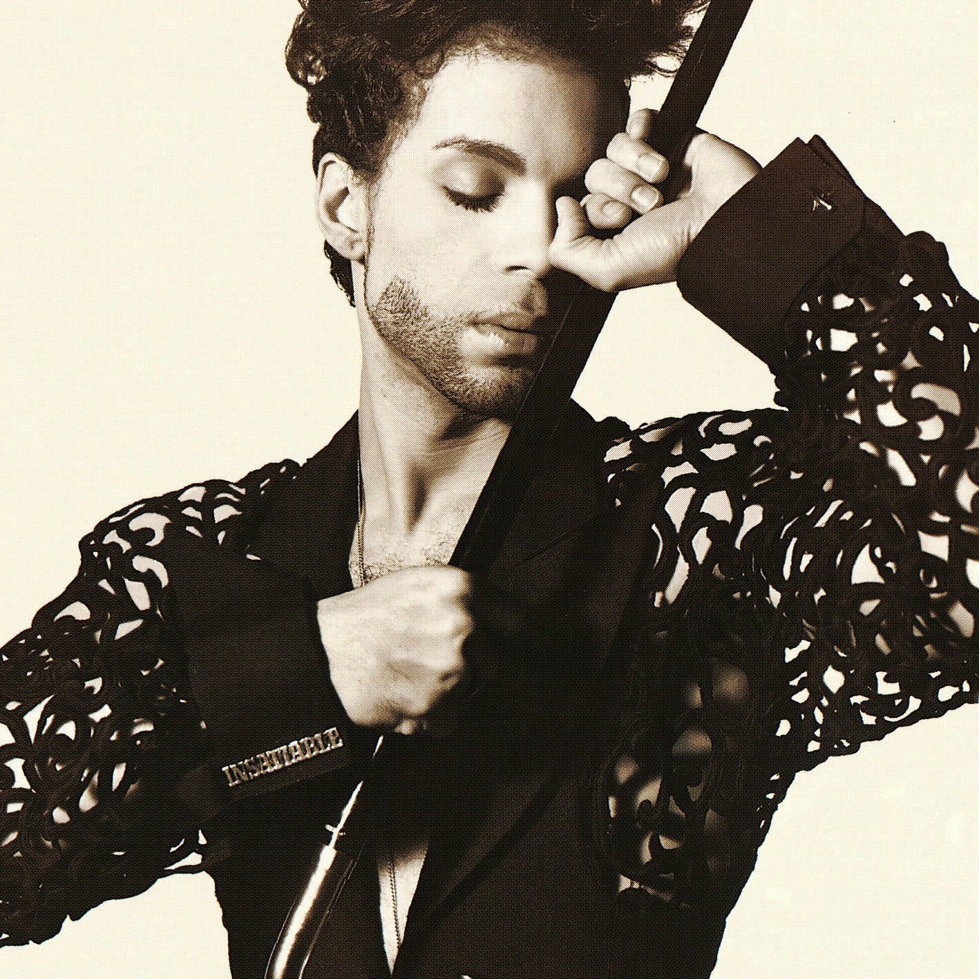 A person with curly hair and a trimmed beard poses with eyes closed, holding a cane and wearing a detailed, cut-out sleeve jacket.