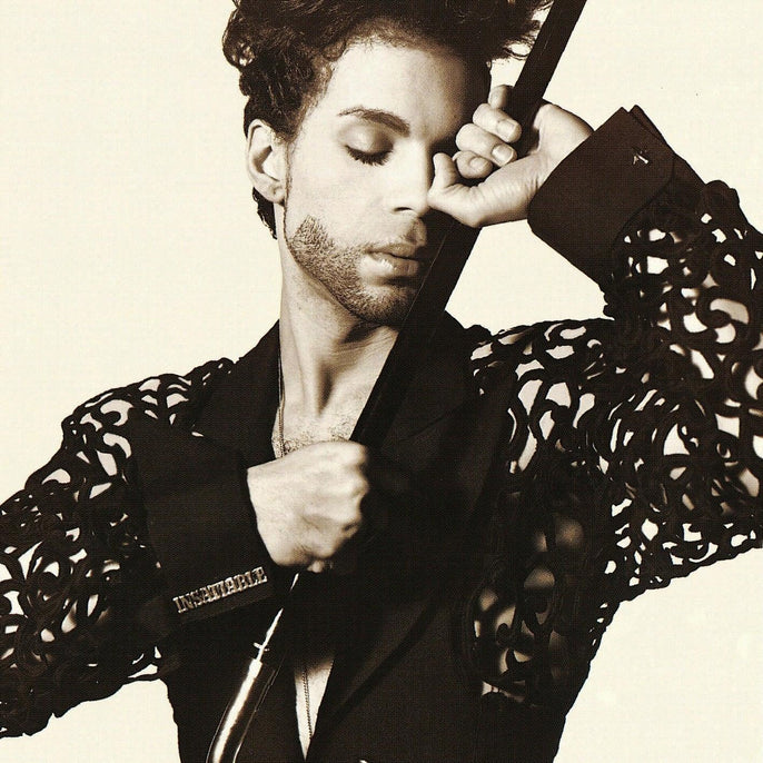 A person with curly hair and a trimmed beard poses with eyes closed, holding a cane and wearing a detailed, cut-out sleeve jacket.