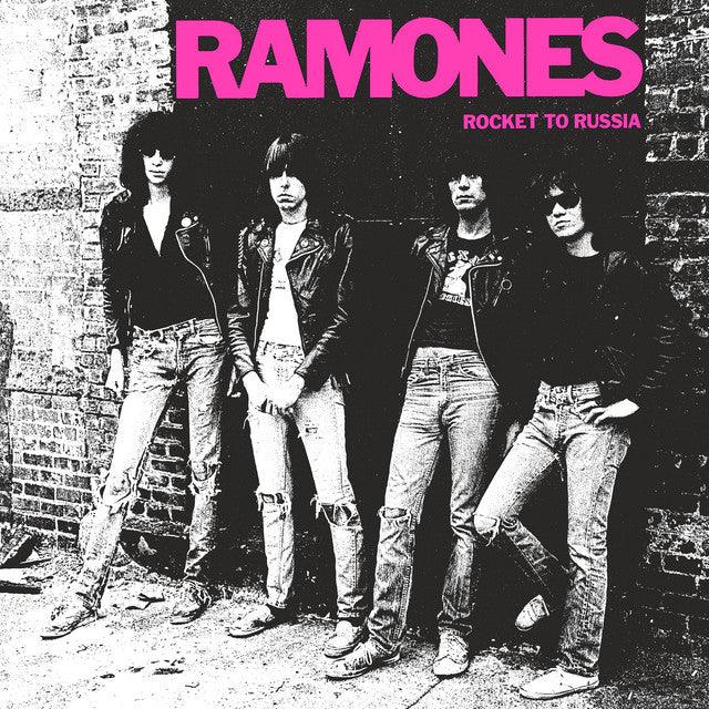 Four men in leather jackets and ripped jeans stand against a brick wall with "RAMONES" and "ROCKET TO RUSSIA" in pink text above them. Black and white photo.
