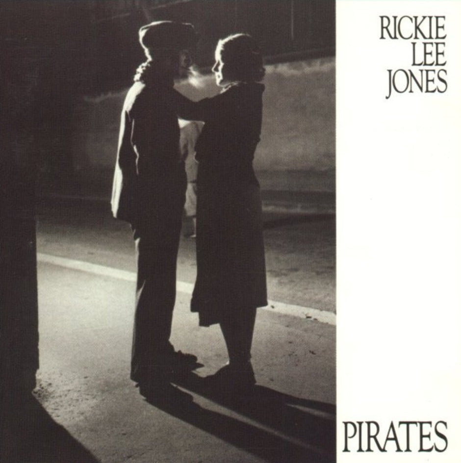 Two people stand facing each other on a dimly lit street at night. The image appears on the cover of Rickie Lee Jones's album titled "Pirates.