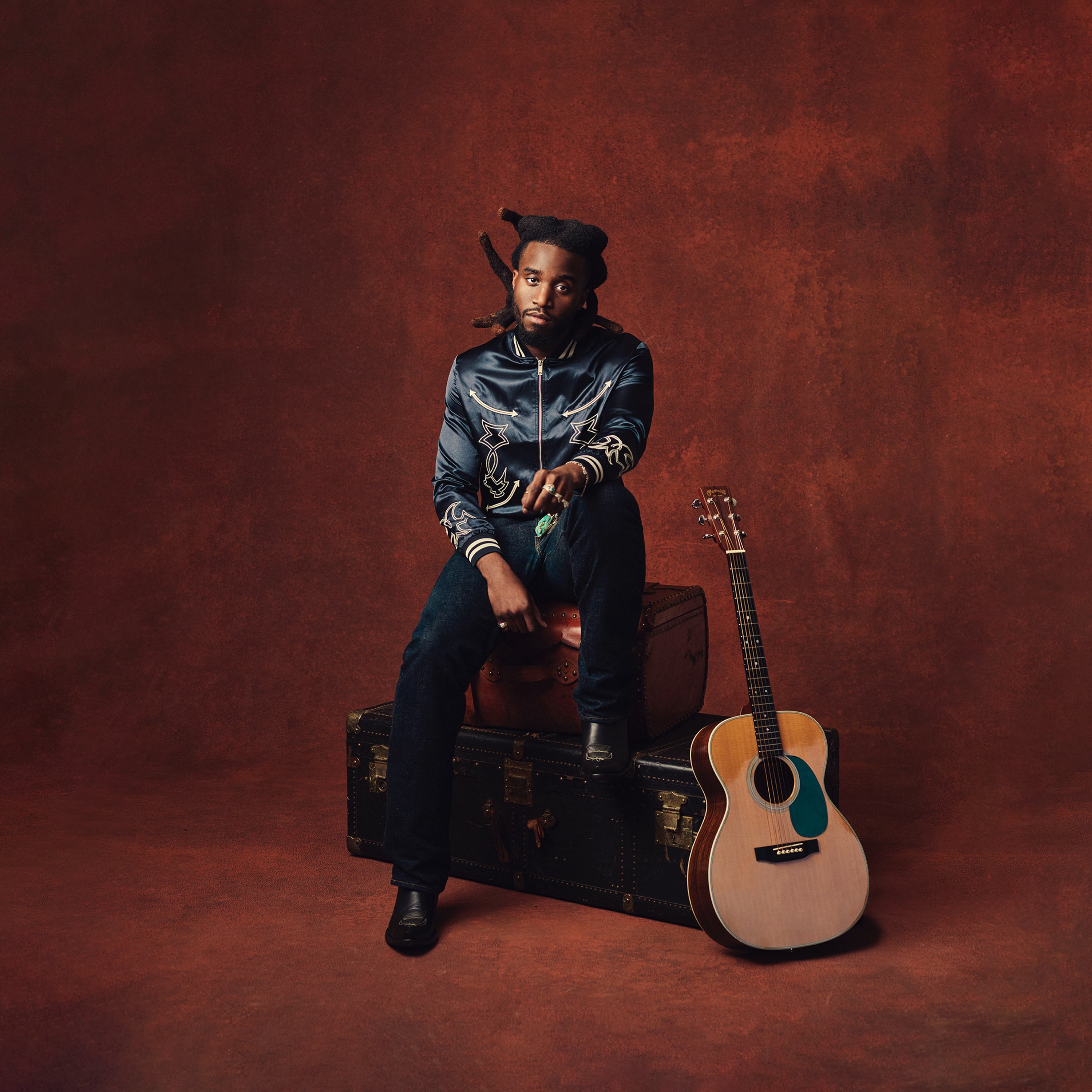 A man with long hair sits on stacked trunks against a reddish-brown backdrop. An acoustic guitar rests upright on the floor beside him.