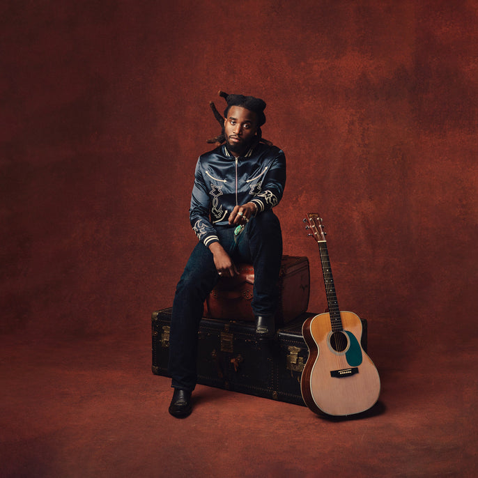 A man with long hair sits on stacked trunks against a reddish-brown backdrop. An acoustic guitar rests upright on the floor beside him.