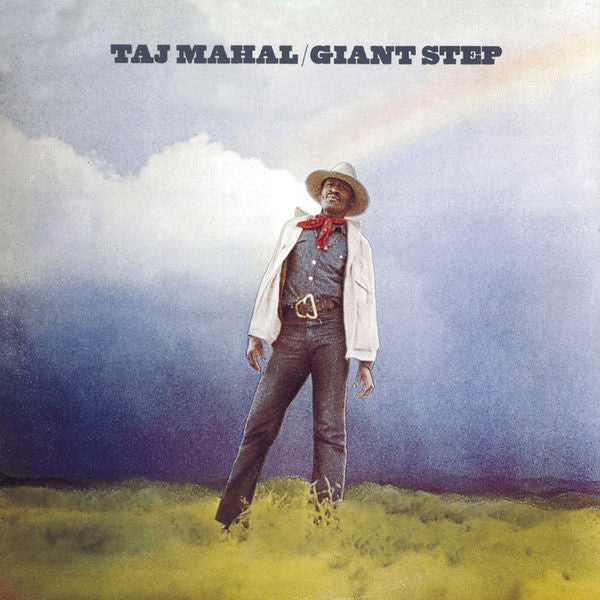 A person wearing a hat and light jacket stands in a grassy field under a cloudy sky with a rainbow; text reads "TAJ MAHAL / GIANT STEP" at the top.