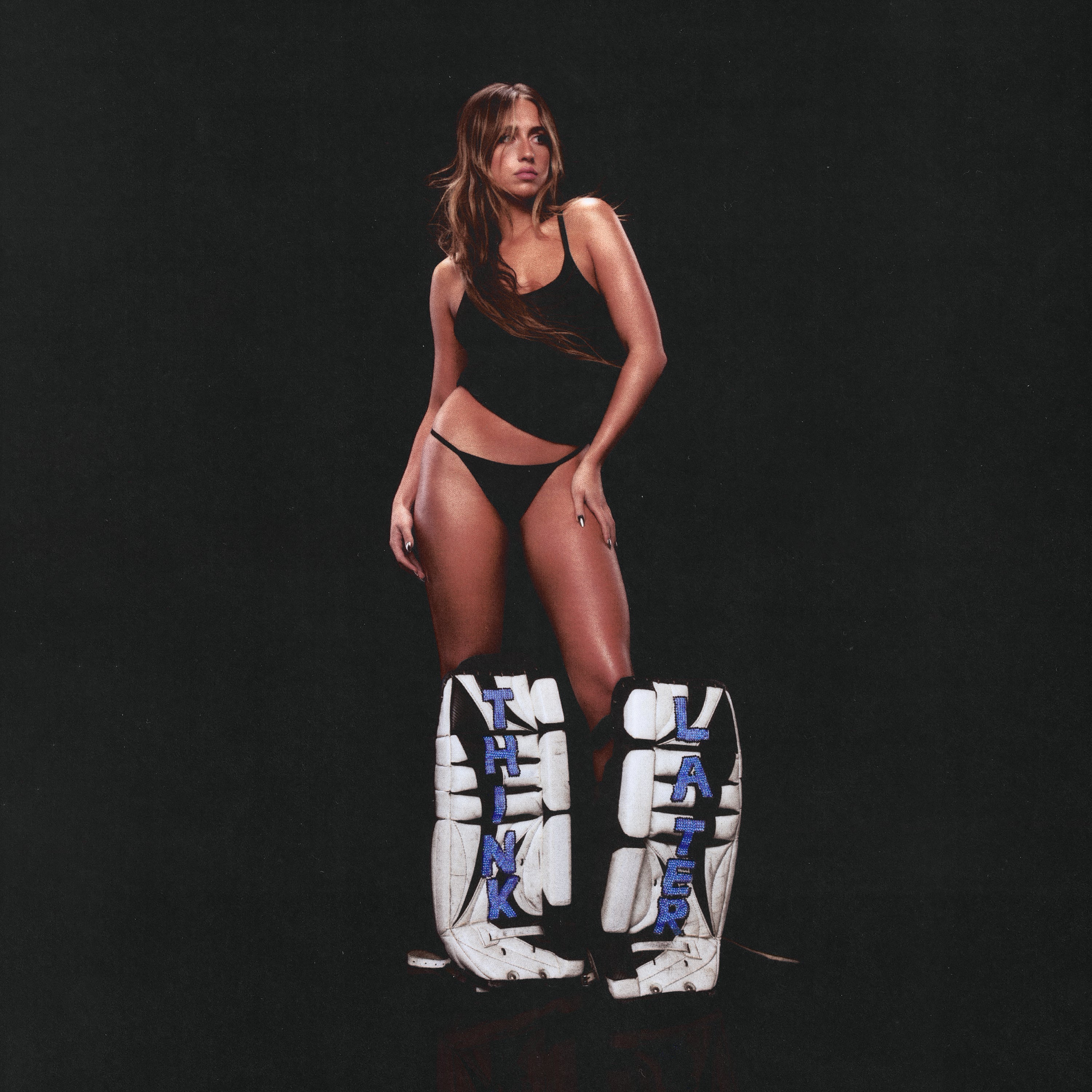 A woman in a black tank top and underwear stands with her hand on her hip behind large white hockey goalie pads labeled "THINK" and "LATER" against a plain black background.