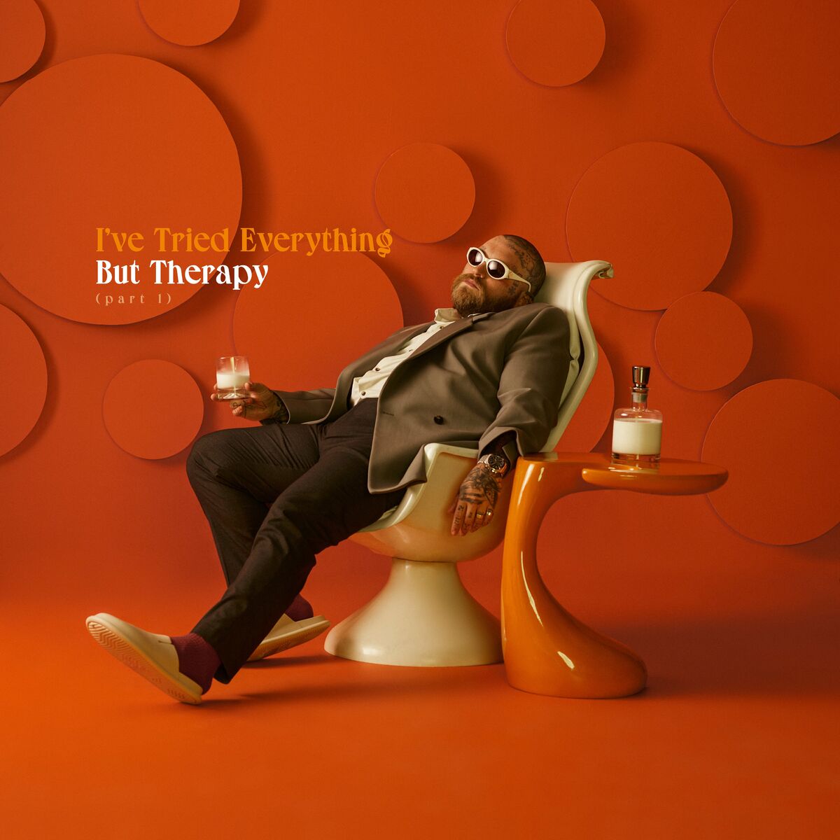 A man in a suit and sunglasses sits in a modern white chair holding a drink, with an orange background and table; text reads "I've Tried Everything But Therapy (Part 1).