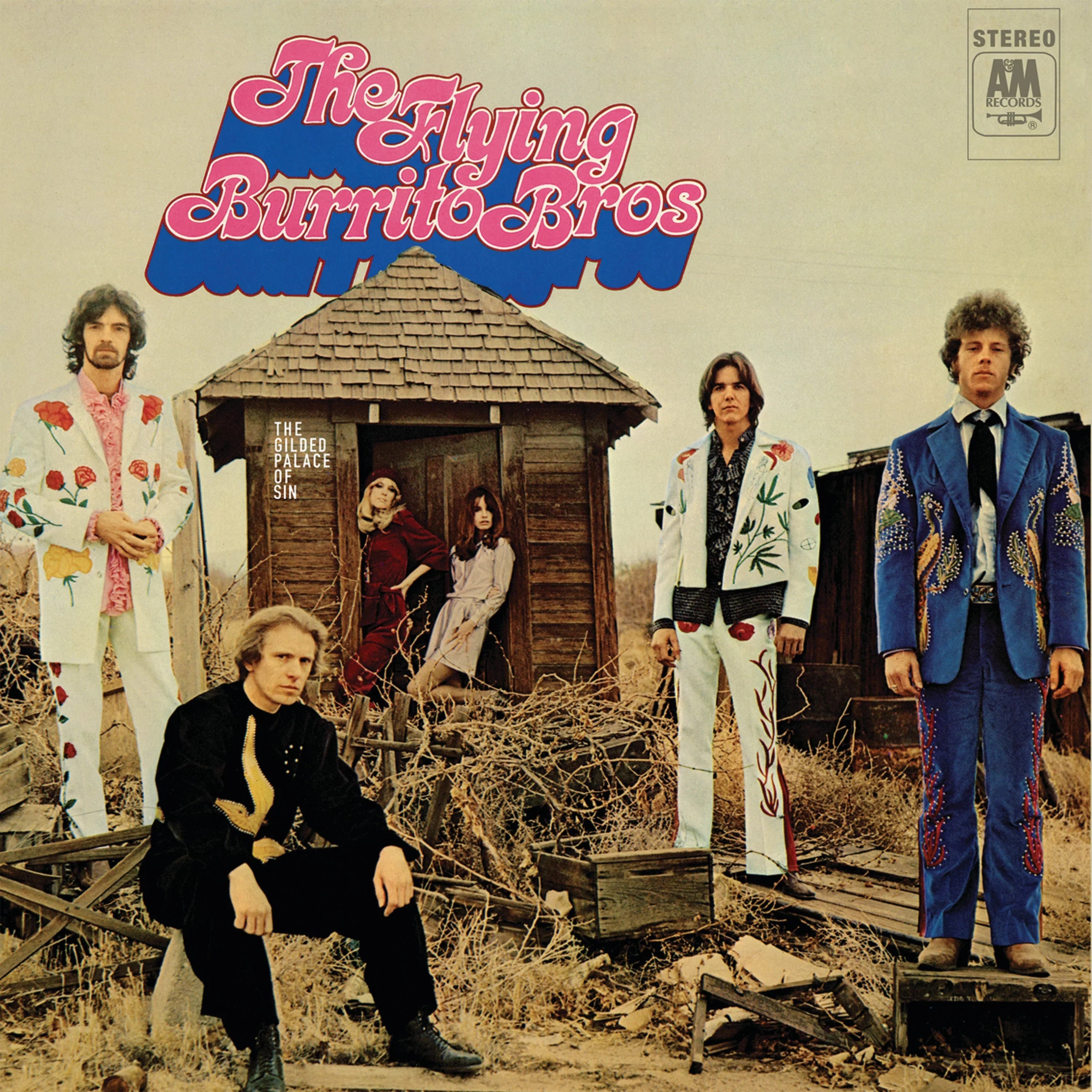 Five men in embroidered suits stand and sit around weathered wooden structures in a dry, outdoor setting under the title "The Flying Burrito Bros.