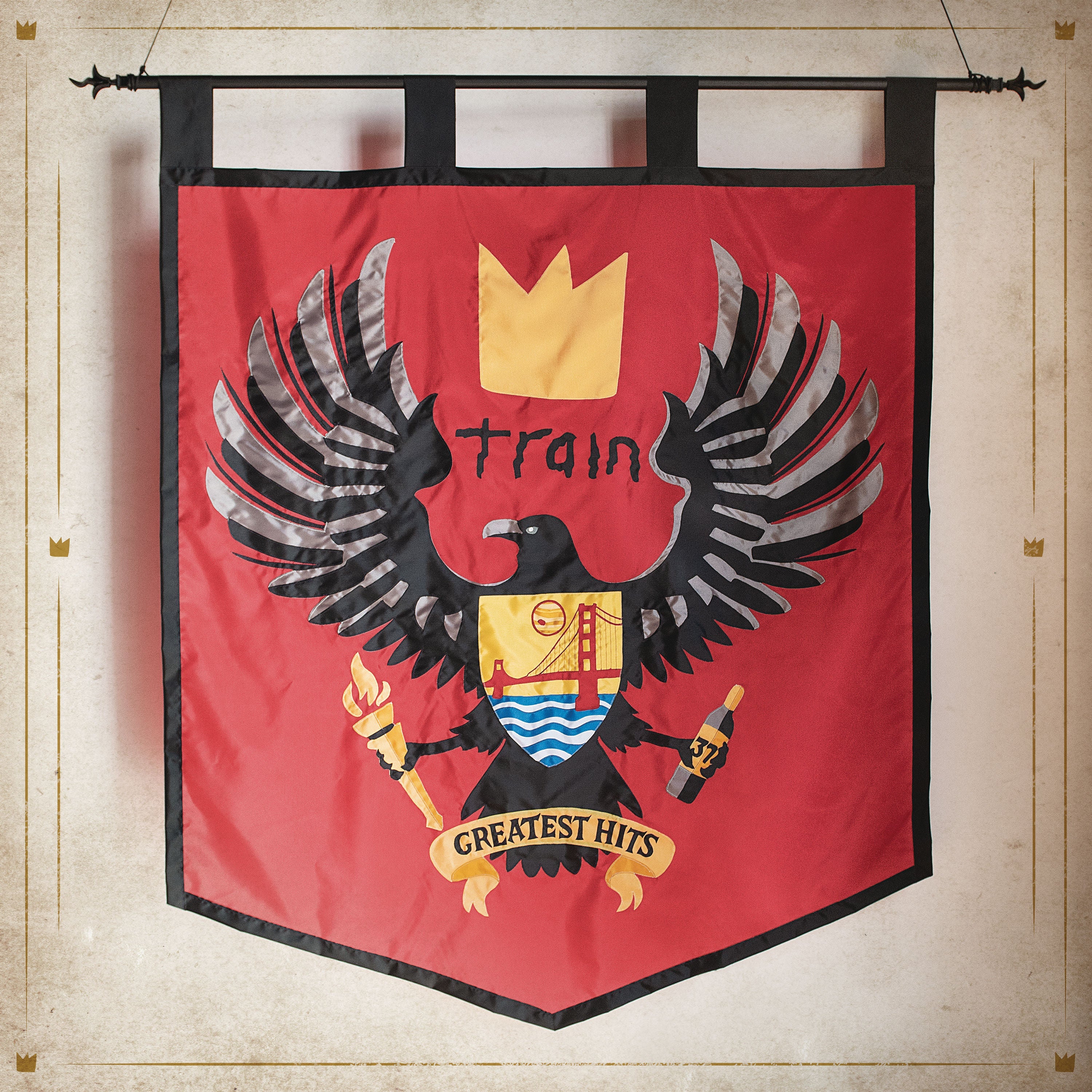A red banner features a black eagle with outstretched wings, a yellow crown, and a shield with various symbols. The word "train" and the text "Greatest Hits" are displayed.