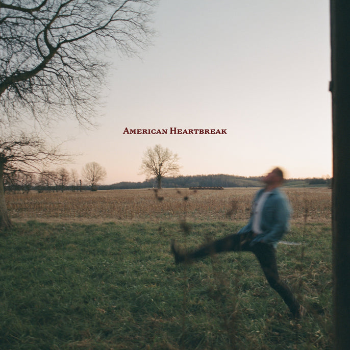 A blurred person mid-kick in a grassy field at dusk with bare trees; text "American Heartbreak" appears in the sky near the top center.