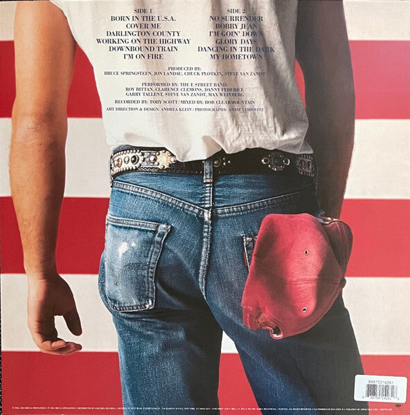Person in blue jeans with red cap in pocket, white shirt with song list, American flag background