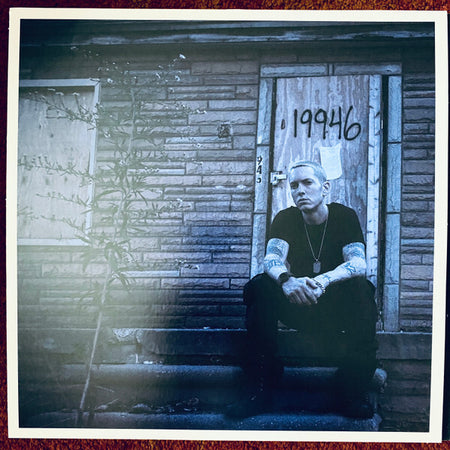 Tattooed man sitting on porch steps in front of a boarded house with street number 19946