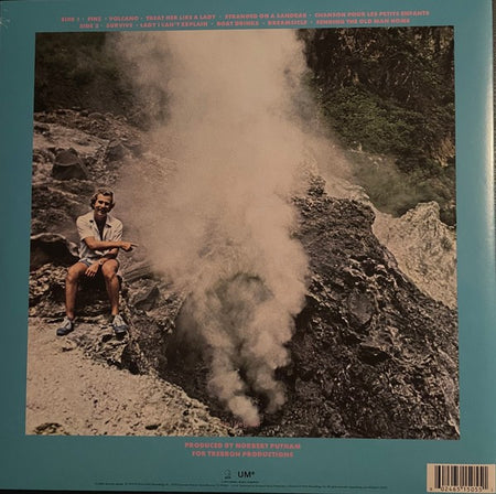 Man sitting near a steaming volcano vent with rocky terrain and greenery in background
