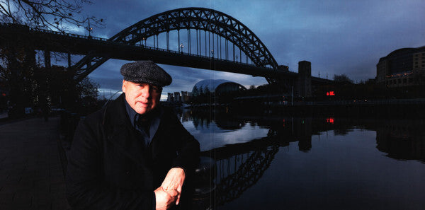 Man in dark coat and flat cap standing by river at dusk with arched bridge in background