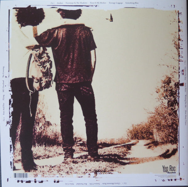 Two people with vintage clothing stand arm in arm on a rural path, holding a guitar, sepia tone.