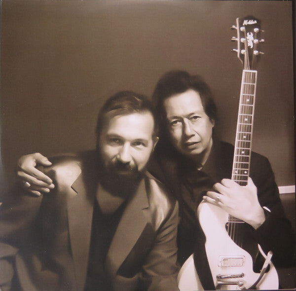 Two men posing together, one holding an electric guitar, in a vintage-style photo