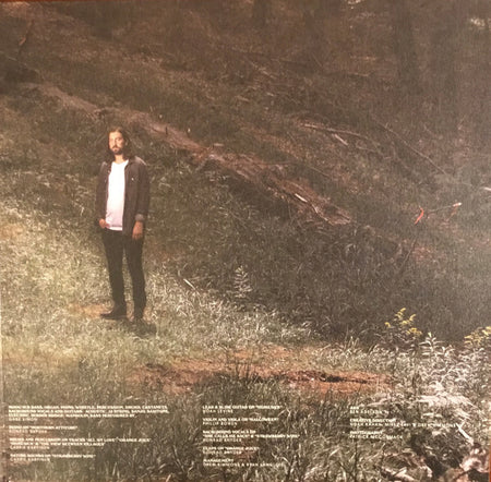 Man standing in a sunlit forest clearing, surrounded by grass and trees, with album credits visible.
