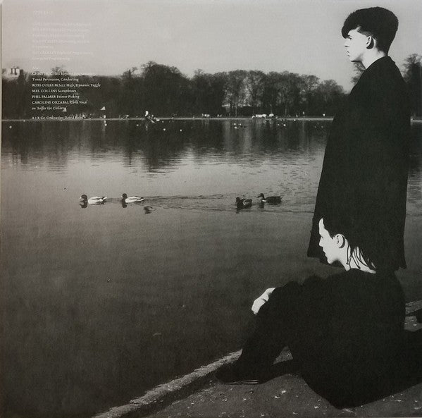 Black and white photo of two people by a lakeside with ducks, trees, and reflections in the water.