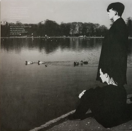 Black and white photo of two people by a lakeside with ducks, trees, and reflections in the water.