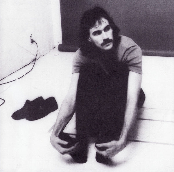 Black and white photo of a man with a mustache sitting on the floor, shoes beside him, minimalist studio setting.
