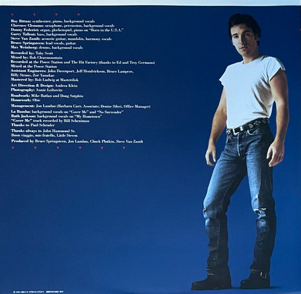 Man in white t-shirt and blue jeans standing against a blue background with album credits text.