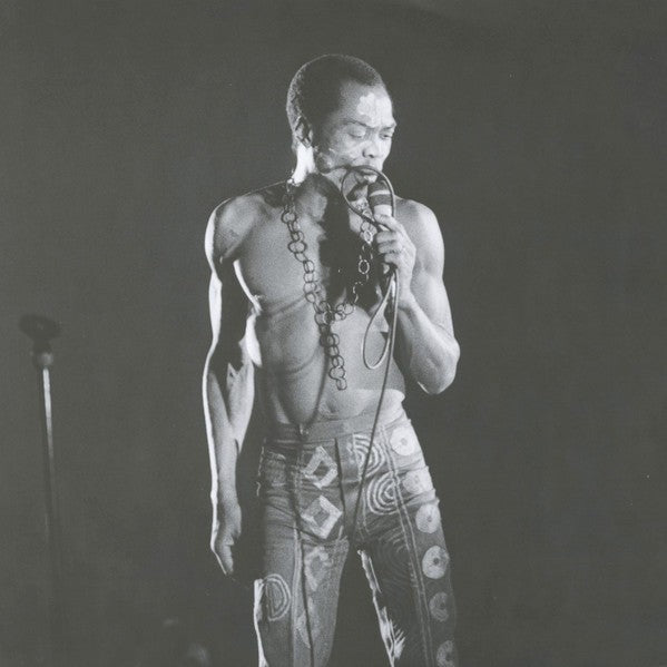 Black and white photo of a shirtless man singing on stage in patterned pants, holding a microphone