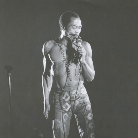 Black and white photo of a shirtless man singing on stage in patterned pants, holding a microphone