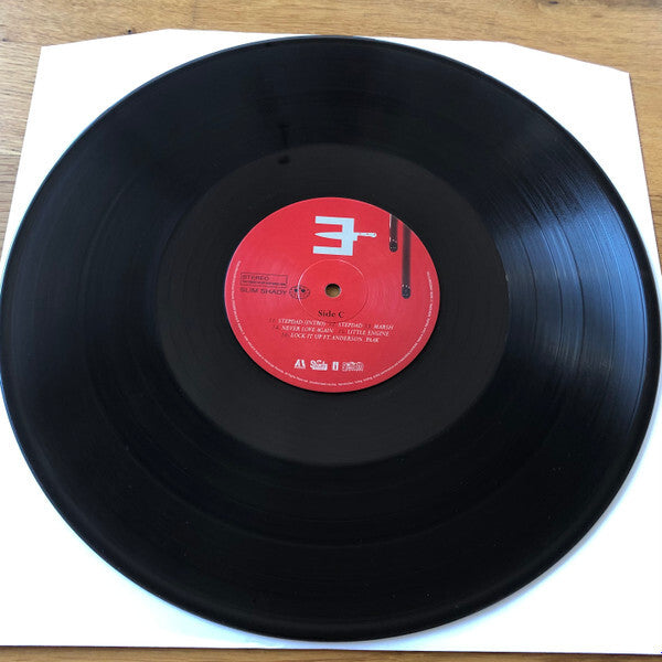 Black vinyl record with a red label on a white sleeve, viewed from above on a wooden surface.