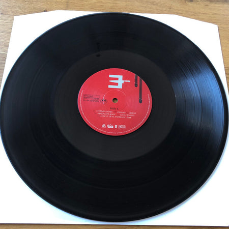 Black vinyl record with a red label on a white sleeve, viewed from above on a wooden surface.